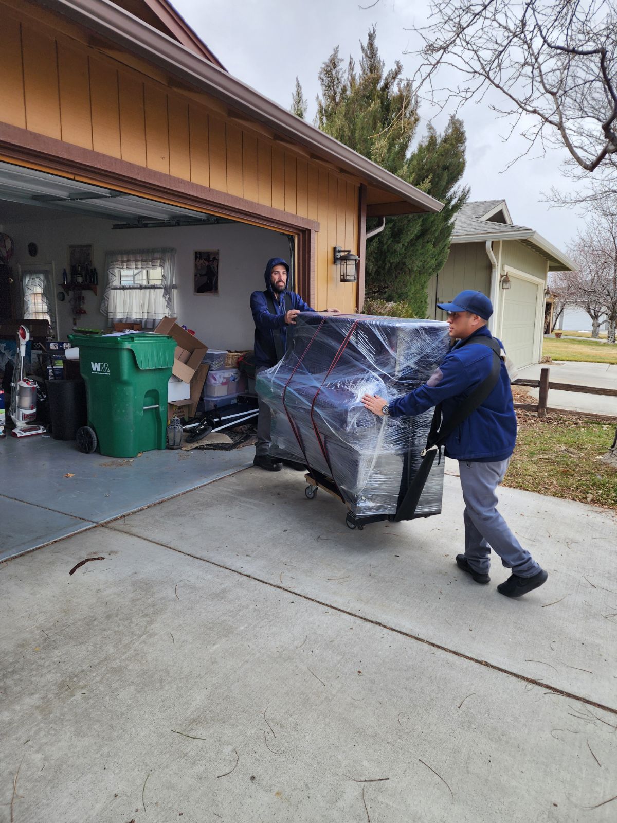 Two Green Echo movers loading a shrink-wrapped piece of furniture from a garage onto a dolly